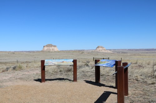 Two informational signs in a barren landscape with distant rock formations under a clear blue sky.