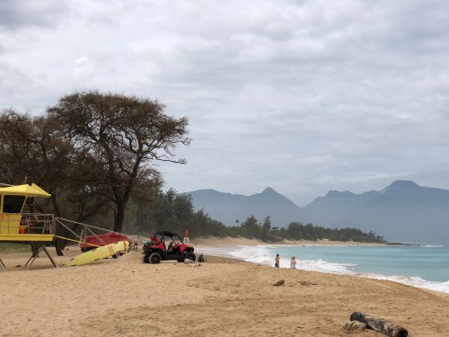 A sandy beach with a lifeguard tower, rescue equipment, and people walking along the shore under a cloudy sky.
