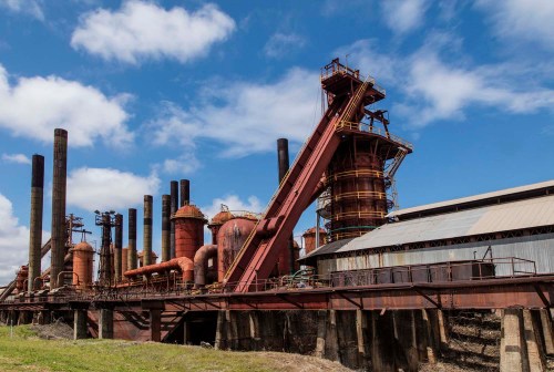 An industrial site with large rusted structures, smokestacks, and a blue sky with scattered clouds.