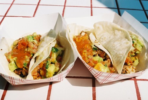Two tacos in paper trays, filled with diced tomatoes, avocado, cheese, and sauce, on a checkered table.