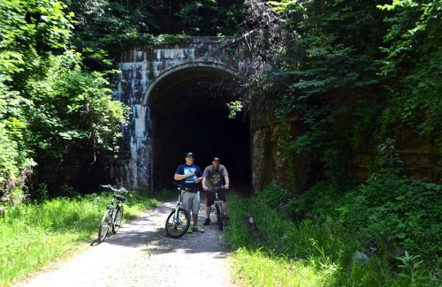 Two cyclists stand near a tunnel entrance surrounded by lush greenery, with their bikes resting beside them.