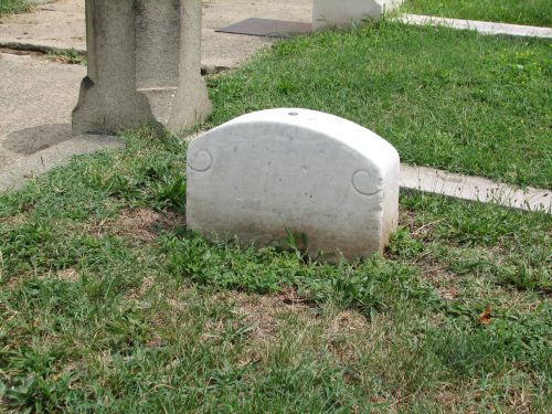 A small, rounded white gravestone partially covered by grass in a cemetery.
