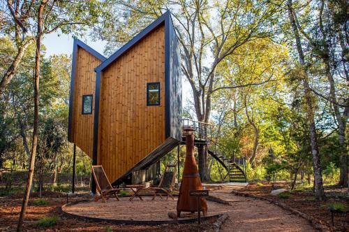 A modern wooden treehouse with a unique design, surrounded by trees and outdoor seating in a natural setting.