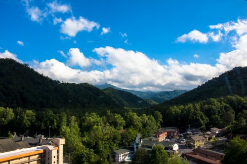 A scenic view of green mountains under a blue sky with fluffy clouds, overlooking a small town below.