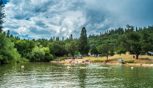 A serene lake scene with people swimming and relaxing on the shore, surrounded by lush green trees and cloudy skies.