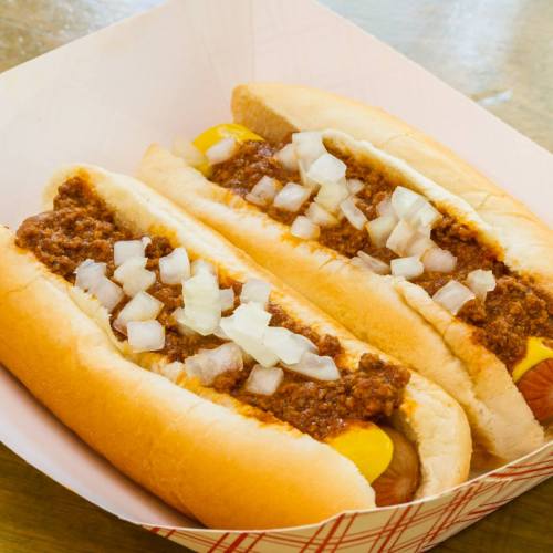 Two hot dogs in buns topped with chili and diced onions, served in a paper tray.