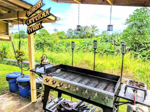Outdoor camp kitchen with a large grill, kettle, and signs for bathroom, camp kitchen, and TV room, surrounded by greenery.
