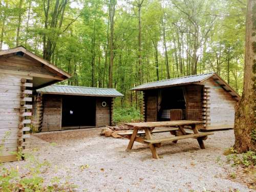 Two wooden cabins in a forested area, with a picnic table in front and gravel ground. Lush green trees surround the scene.