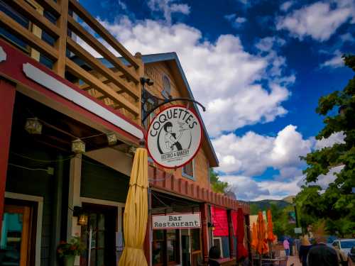 Sign for Coquettes Bistro & Bar, featuring outdoor seating and a colorful building under a blue sky with clouds.