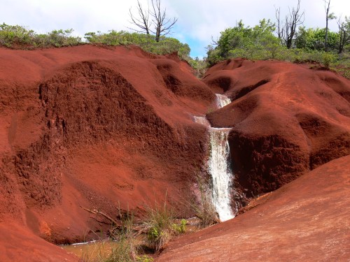 A vibrant red landscape with a small waterfall cascading down rocky terrain, surrounded by green vegetation.