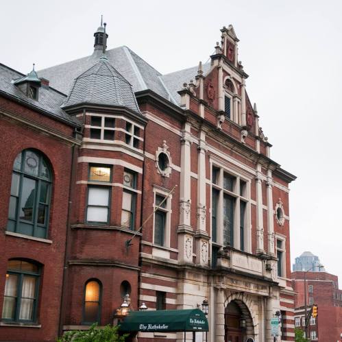 Historic brick building with ornate architecture, featuring a peaked roof and large windows, set against a cloudy sky.