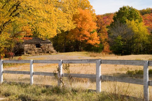 A rustic stone building surrounded by vibrant autumn trees and a white wooden fence in a serene landscape.