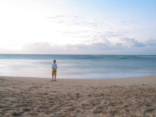 A child stands on a sandy beach, gazing at the calm ocean under a cloudy sky.