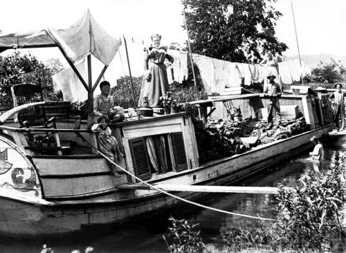 A historic black-and-white photo of a barge with people, laundry hanging, and greenery along a waterway.