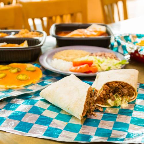 A table with various Mexican dishes, including a burrito, rice, beans, and enchiladas on colorful checkered tablecloth.