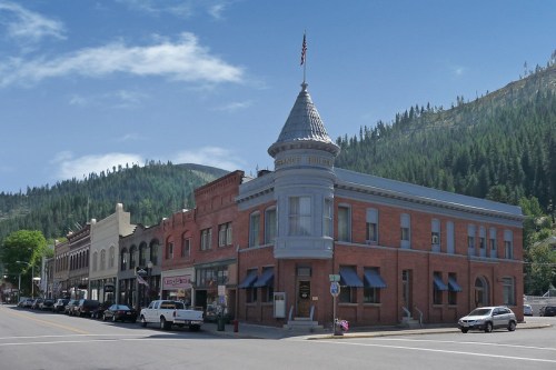 Historic buildings line a street with a prominent towered structure, set against a backdrop of green mountains.