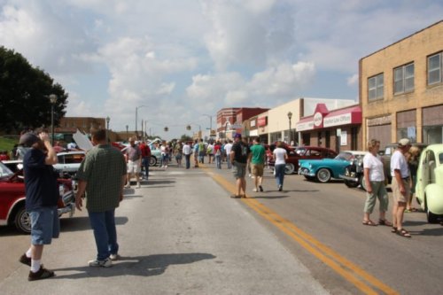 A bustling street filled with people admiring classic cars during a car show, with shops lining the road.