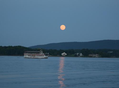 A riverboat glides on calm water under a large, glowing full moon against a twilight sky and distant hills.
