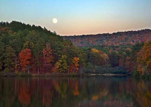 A serene lake reflects colorful autumn trees and a full moon against a twilight sky.