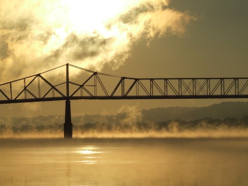 A silhouette of a bridge over a misty river at sunrise, with clouds and sunlight creating a serene atmosphere.