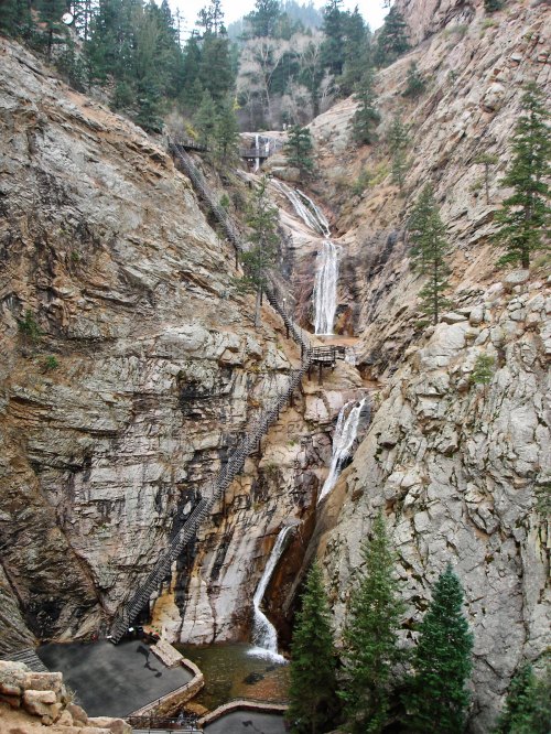 A scenic view of a waterfall cascading down rocky cliffs, surrounded by pine trees and rugged terrain.