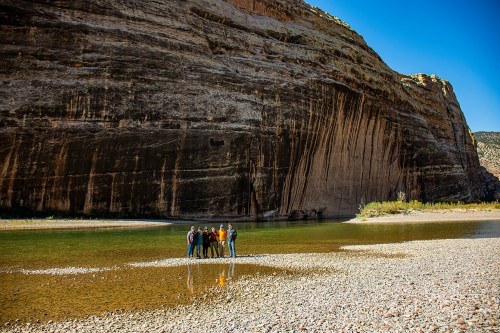 A group of people stands on a rocky riverbank with a large cliff in the background under a clear blue sky.
