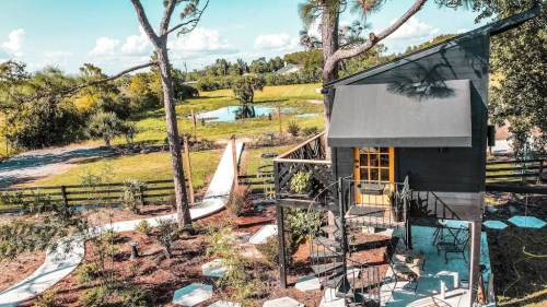 A cozy cabin surrounded by greenery, featuring a spiral staircase and outdoor seating, with a pond in the background.