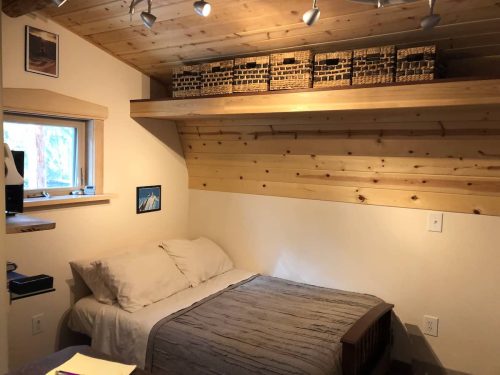 Cozy bedroom with a wooden ceiling, a bed, and shelves holding woven baskets. Natural light from a window.