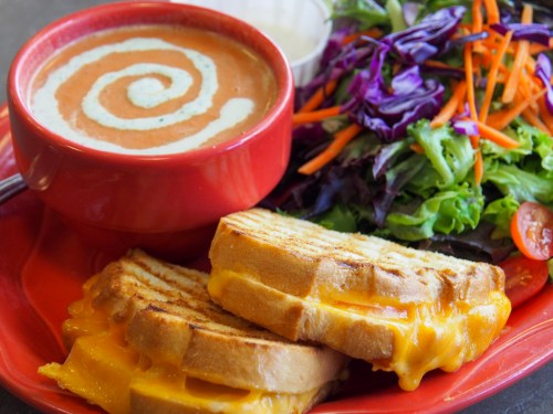A bowl of tomato soup with a swirl of cream, served with a side salad and two grilled cheese sandwiches.