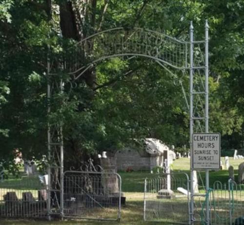 A wrought iron archway marks the entrance to a cemetery, surrounded by trees and a sign indicating visiting hours.