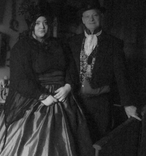 A man and woman in vintage attire pose together in a dimly lit setting, with a classic, elegant style.