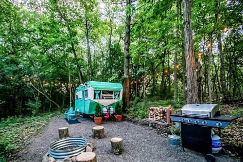 A vintage teal camper surrounded by trees, with a grill, fire pit, and string lights creating a cozy outdoor setting.