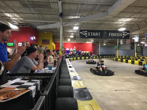 A go-kart track with racers speeding by, while spectators watch from the sidelines, cheering and taking photos.