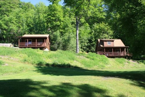 Two rustic cabins nestled among lush green trees, surrounded by a grassy area and a serene landscape.