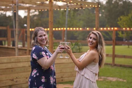 Two women toast with glasses of red drinks, smiling in a festive outdoor setting with string lights in the background.