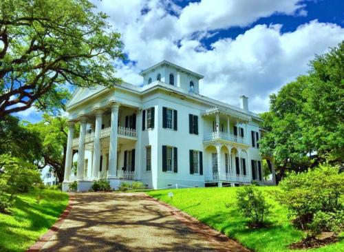 A grand white mansion with columns, surrounded by lush greenery and a blue sky with fluffy clouds.