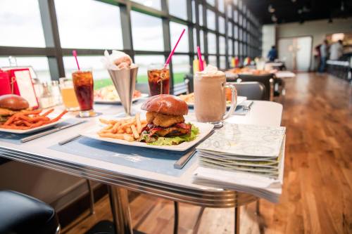 A table set with a burger, fries, sweet potato fries, drinks, and a milkshake in a modern restaurant with large windows.