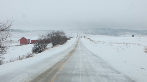 A snowy rural road stretches into the distance, flanked by bare trees and a red barn under a gray, overcast sky.