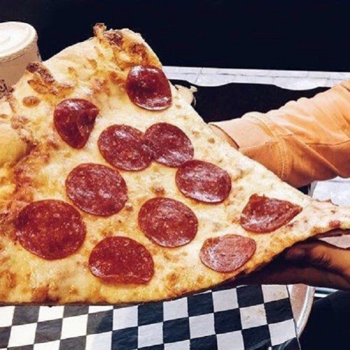 A large slice of pepperoni pizza held by a hand, with a crispy crust and melted cheese on a checkered table.