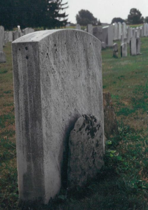 A weathered gravestone in a cemetery, partially obscured by grass, with other gravestones visible in the background.