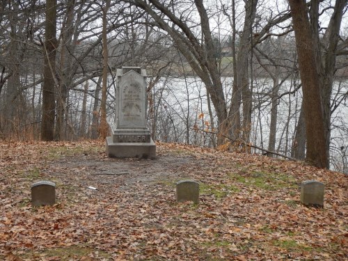 A weathered gravestone surrounded by bare trees and fallen leaves, near a riverbank.
