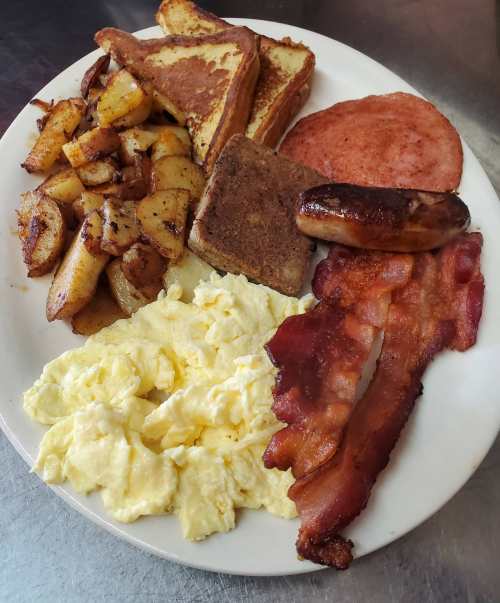 A hearty breakfast plate with scrambled eggs, bacon, sausage, ham, French toast, and seasoned potatoes.