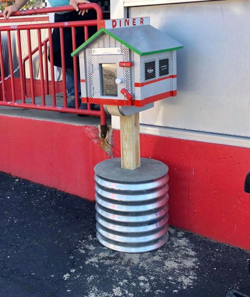 A whimsical diner-themed mailbox on a metal post, featuring a colorful house design and a red railing in the background.