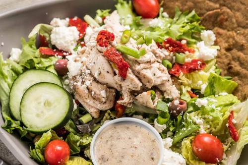 A fresh salad with grilled chicken, cucumbers, cherry tomatoes, olives, feta cheese, and a side of dressing.