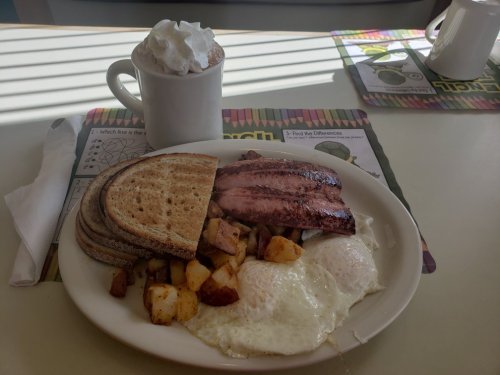 A plate with two eggs, roasted potatoes, slices of toast, and a side of bacon, with a hot chocolate in a mug.