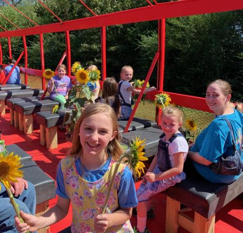 A group of children and adults on a red wagon, holding sunflowers and enjoying a sunny day outdoors.