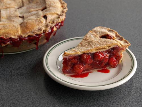 A slice of cherry pie on a plate, with the whole pie in the background, oozing with cherry filling.
