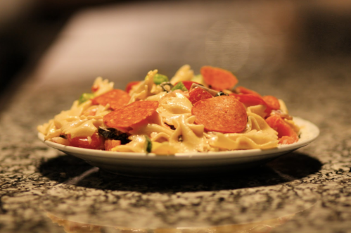 A plate of pasta topped with pepperoni slices, tomatoes, and green vegetables on a textured surface.