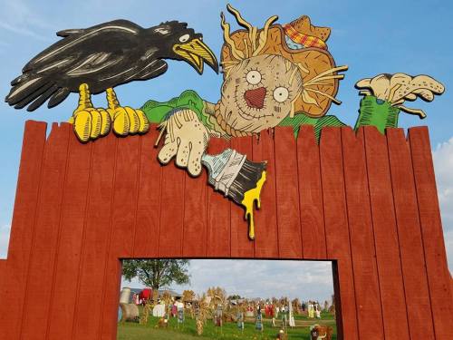 A colorful scarecrow and crow cutout atop a wooden fence, with a field of hay bales and decorations in the background.