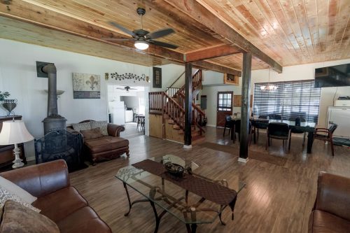 Cozy living room with wooden beams, a staircase, and a dining area, featuring leather sofas and a glass coffee table.